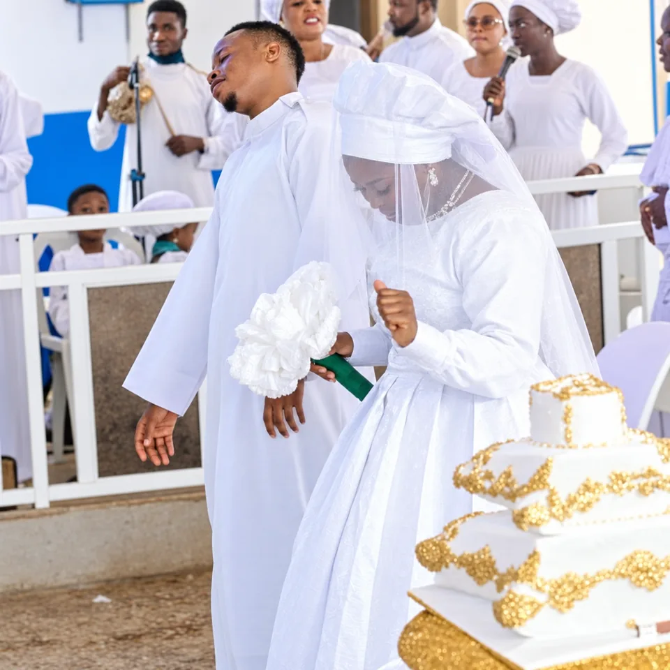 A bride and groom stand over a tiered wedding cake iced in white and gold, the choir behind them