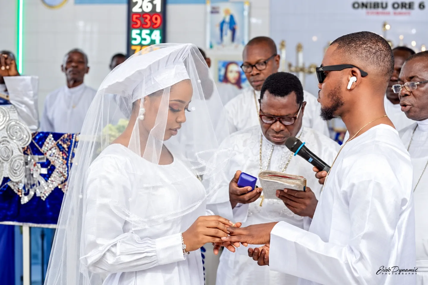 A groom places a ring on a bride's finger while a minister holds an open Bible beside them